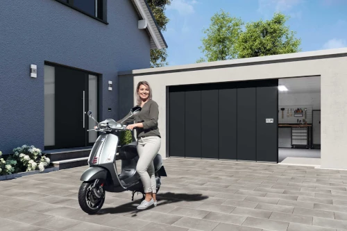 Frau steht mit Motorroller vor moderner Garage mit geöffnetem Sektionaltor und Einfamilienhaus.
