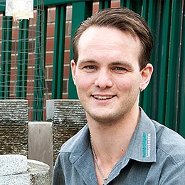 Junger Mann mit grauem Hemd lächelt vor Brunnen und Gartenelementen im Außenbereich.