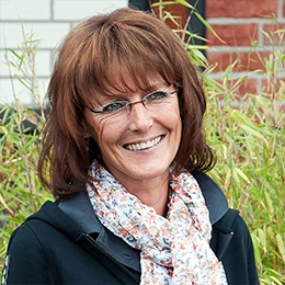 Frau mit Brille und floralem Schal lächelt vor grünen Pflanzen im Außenbereich.