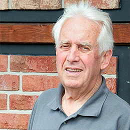 Älterer Mann mit grauen Haaren steht vor einer Backsteinwand und trägt ein graues Polohemd.