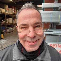Ein lächelnder Mann mit Brille steht in einem Baustofflager mit Regalen und Baumaterialien.