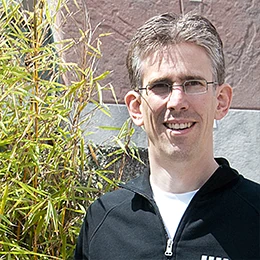 Ein Mann mit Brille steht draußen vor einer Steinwand und Bambuspflanzen im Sonnenlicht.