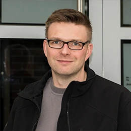 Mann mit Brille und dunkler Jacke steht vor einer modernen Eingangstür und lächelt.