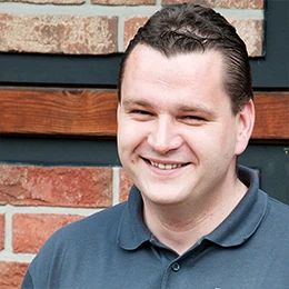 Mann mit dunklen Haaren lächelt vor einer Backsteinwand und trägt ein dunkelblaues Polohemd.