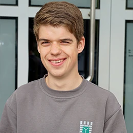 Junger Mann mit braunen Haaren lächelt vor einem modernen Gebäude mit Glastüren.