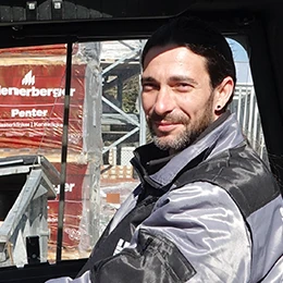 Ein Bauarbeiter sitzt in Arbeitskleidung auf einer Baustelle mit Baumaterial im Hintergrund.